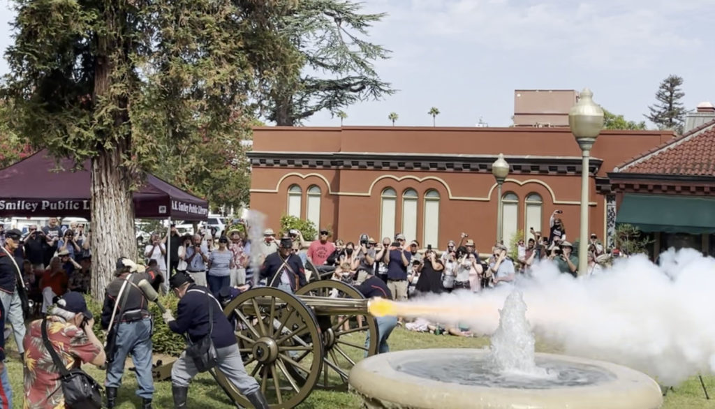 Photograph of the Lincoln Shrine's reproduction cannon being fired in 2022. Photo shows a bronze cannon on a wooden carriage being fired with a group of people in the background. 